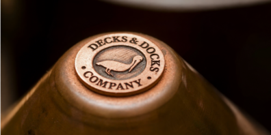 Close-up of a copper Decks & Docks Company medallion, featuring a wading bird icon, embossed on the wooden base of a dark, tapered mug or bowl.